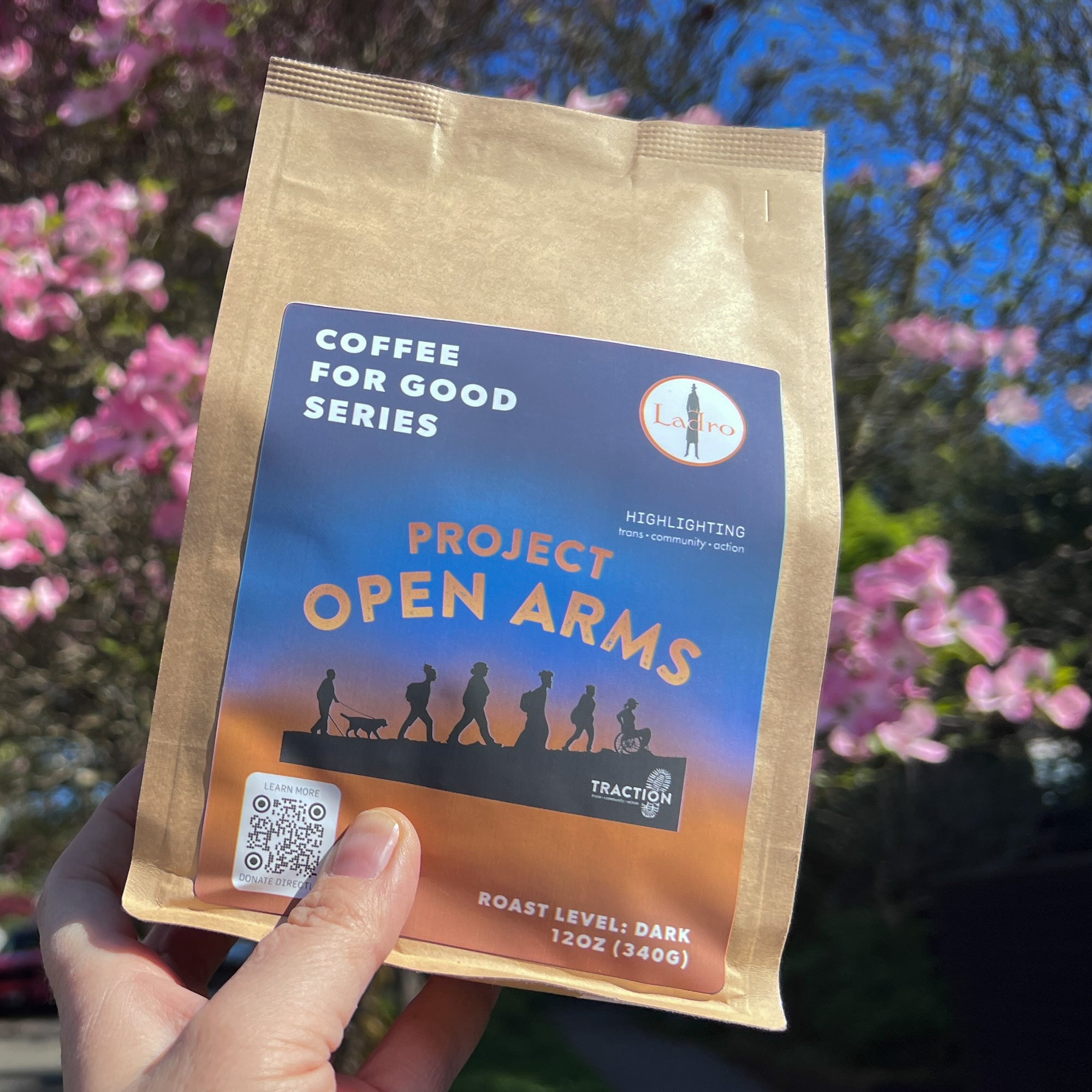 Coffee package labeled 'Project Open Arms' held in front of a blurred natural background with pink flowers.